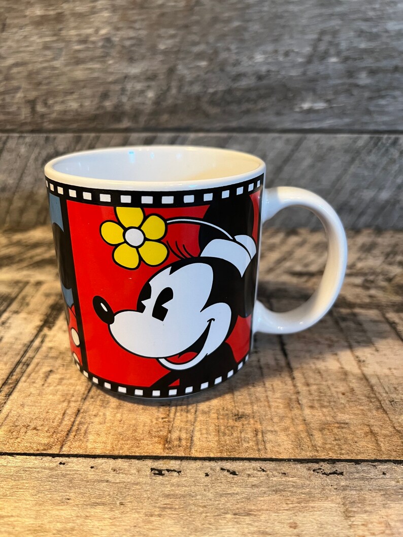 May include: A white ceramic mug with a black and red design featuring Mickey Mouse and Minnie Mouse. The design includes a yellow flower and a film strip border.