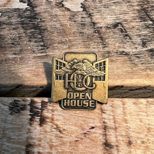 May include: A rectangular, gold-colored pin with the text "OPEN HOUSE" at the bottom. The pin features an eagle with outstretched wings above the letters "HOC". The pin is set against a wooden background.