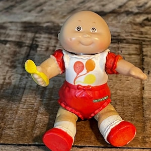 May include: A vintage Cabbage Patch Kids doll with a bald head, smiling face, and brown eyes. The doll wears a red outfit with a white bib featuring balloon graphics and holds a yellow spoon. The doll's red shoes and white socks complete the look.