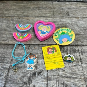 May include: A collection of Cabbage Patch Kids items including two rainbow-shaped erasers, a pink heart-shaped case with a doll illustration, a yellow circular pin, a blue cord necklace with a doll pendant, a yellow alphabet card, and a small pin.