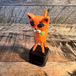 May include: Orange cat figurine with blue eyes, black accents, and a green bow. The cat is standing on a black rectangular base. The cat's tail is upright. The figurine is made of plastic.