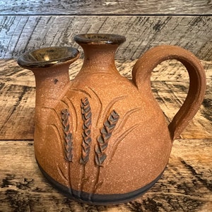 May include: A brown ceramic pitcher with a handle and two spouts. The pitcher has a textured surface and a design of wheat stalks.