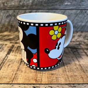 May include: A white ceramic mug with a black and white film strip border. The mug features a cartoon image of Mickey Mouse and Minnie Mouse. Mickey is on the left side of the mug and Minnie is on the right. Mickey is wearing red shorts and Minnie is wearing a red dress with yellow flowers.