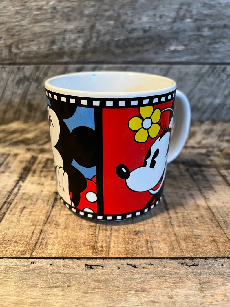May include: A white ceramic mug with a black and white film strip border. The mug features a cartoon image of Mickey Mouse and Minnie Mouse. Mickey is on the left side of the mug and Minnie is on the right. Mickey is wearing red shorts and Minnie is wearing a red dress with yellow flowers.