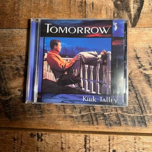May include: A music CD case titled Tomorrow by Kirk Talley. The cover art features a person in an orange shirt and light-colored pants sitting on a white railing at dusk. The text Tomorrow is at the top, and Kirk Talley is at the bottom.