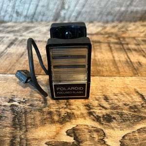 May include: A vintage Polaroid Focused Flash unit. The black flash has a rectangular shape with a silver-toned grid and the text "POLAROID FOCUSED FLASH". A black cord and clip are attached to the flash.