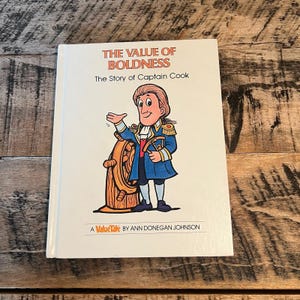 May include: A children's book titled "The Value of Boldness" featuring a cartoon illustration of Captain Cook. The cover has the text "The Story of Captain Cook" and "A ValueTale by Ann Donegan Johnson". The book is on a wooden surface.