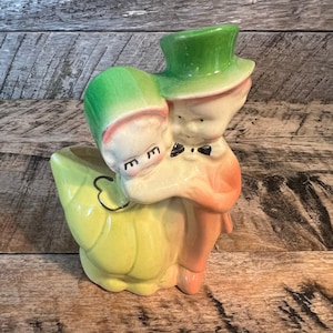 May include: A vintage ceramic planter in the shape of a couple embracing. The figures are painted with a cream base, with green hats, and peach-colored clothing. The planter has a yellow and green leaf-shaped base.