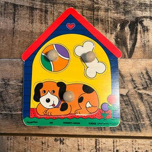 May include: A wooden puzzle in the shape of a dog house. The puzzle features a cartoon dog, a ball, and a bone. The roof is red, the walls are blue, and the interior is yellow. The puzzle has three wooden knobs.