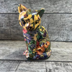 May include: A small ceramic cat figurine in a sitting position, featuring a vibrant, multi-colored floral pattern across its entire surface. The design includes shades of yellow, orange, purple, and green, with dark eyes and a small nose.