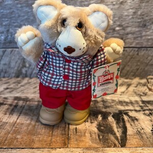May include: A plush bear wearing a red and black plaid shirt, red pants, and tan shoes. The bear has a white snout and ears, and a tag that reads "Wendy's Hamburgers."