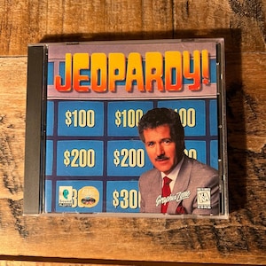 May include: A Jeopardy! PC game jewel case featuring a blue game board grid with dollar amounts and a photograph of Alex Trebek wearing a gray suit and red tie. The cover includes the Sound Blaster logo, Graphix Zone text, and an ESRB KA rating.