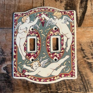 May include: A double light switch plate featuring a decorative illustration of three winged cherubs against a muted teal background. The border is adorned with a pattern of small red and yellow fruits, accented by flowing yellow ribbons.