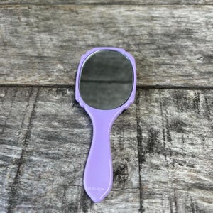 May include: A handheld mirror with a smooth, light purple plastic frame and handle. The mirror features an oval-shaped reflective glass surface set within a scalloped edge border. The handle is solid, rounded, and extends from the base of the frame.