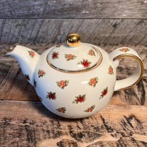 May include: A white teapot with a gold lid and handle. The teapot is decorated with a pattern of small red and yellow roses.