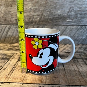 May include: A white ceramic mug with a red, black and yellow design featuring Minnie Mouse. The mug has a black handle and a film strip border.
