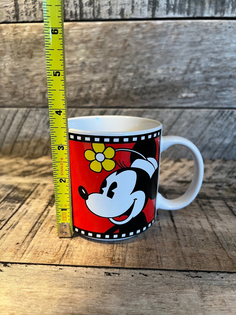 May include: A white ceramic mug with a red, black and yellow design featuring Minnie Mouse. The mug has a black handle and a film strip border.