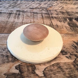 May include: A round, off-white ceramic lid with a light brown wooden knob. The lid has a slightly textured surface and a beveled edge. The knob is a smooth, rounded shape. The lid is likely for a jar or container.