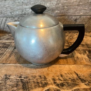 May include: A silver-colored teapot with a black handle and lid knob. The teapot has a rounded body and a spout. The handle is rectangular and curves downward. The lid knob is cone-shaped.