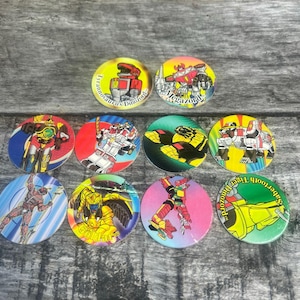 May include: A collection of colorful, round collectible Power Rangers trading game pieces. Each piece features a different Power Ranger character or Zord, with vibrant illustrations and text. The pieces are arranged on a wooden surface.