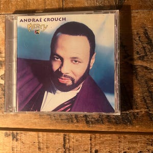 May include: A compact disc case featuring the album Mercy by Andraé Crouch. The cover art displays a portrait of a man with a beard, wearing a dark purple jacket over a white shirt, set against a blue background with the artist and title text.