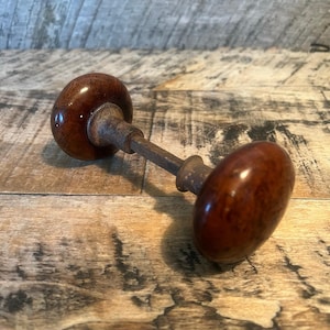 May include: A vintage door knob set featuring two round, polished dark brown wooden knobs connected by a central square metal spindle. The hardware exhibits a weathered, aged patina on the metal components, contrasting with the smooth, glossy finish of the wood. This functional door hardware assembly is designed for installation on a door, showcasing a classic, simple construction with a visible metal shaft connecting the two spherical handles.