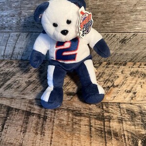May include: A white and blue plush bear wearing a racing uniform with the number 2. The bear has black eyes and a tag that reads "Team Speed Bears". The bear is standing on a wooden surface.