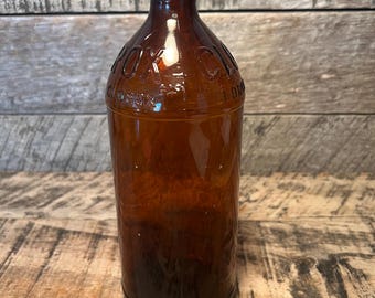 May include: A vintage brown glass bottle with embossed text. The bottle has a long neck and a rounded body. The glass has a dark amber color. The bottle is sitting on a wooden surface.