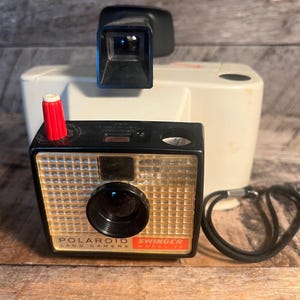 May include: A vintage Polaroid Swinger Land Camera, model 20, in black and beige. The camera has a red button and a black strap. The camera is sitting on a wooden surface.