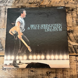 May include: A cassette box set featuring Bruce Springsteen & The E Street Band Live/1975-85. The album cover shows Springsteen holding a guitar on stage, with the band's name and album title in white text. The box set includes 3 cassettes.