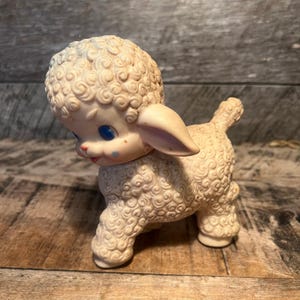 May include: A vintage cream-colored lamb toy with blue eyes, pink accents, and a curly wool texture. The lamb is standing on all fours, with its head tilted down. A classic children's toy.