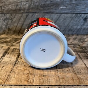 May include: White ceramic mug with a red, black and yellow design featuring Mickey Mouse and other Disney characters. The bottom of the mug reads "&copy; DISNEY JAPAN".