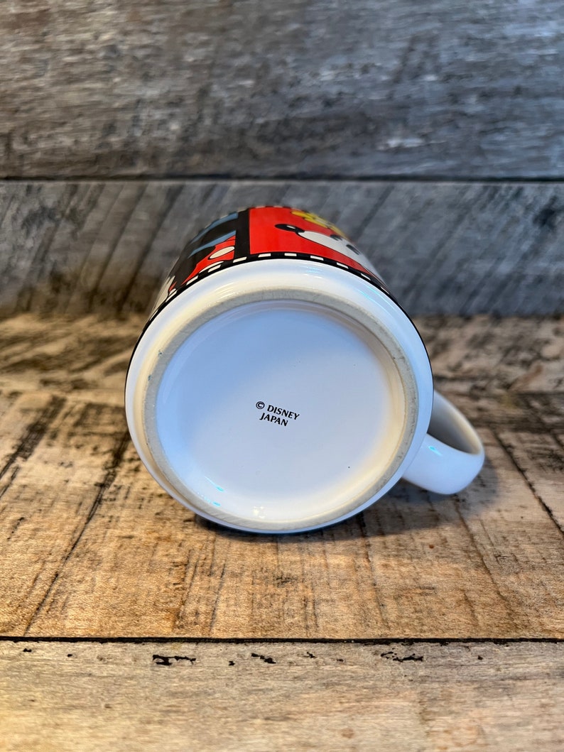 May include: White ceramic mug with a red, black and yellow design featuring Mickey Mouse and other Disney characters. The bottom of the mug reads "&copy; DISNEY JAPAN".