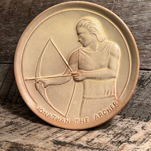 May include: A round, tan-colored ceramic plate featuring a relief of a man with a bow and arrow. The text "JONATHAN THE ARCHER" is inscribed around the edge of the plate. The plate is set against a weathered wooden surface.