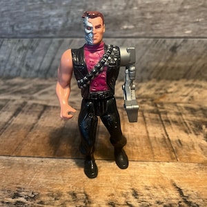 May include: A vintage action figure of a cyborg character. The figure has a human face and hair on one side, and a metallic skull on the other. It wears a black vest, a pink shirt, and black pants. The right arm is a silver robotic arm.
