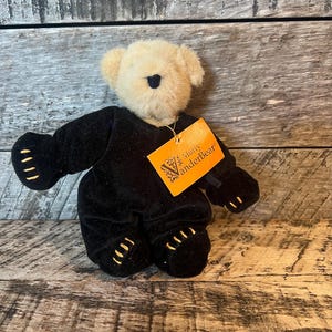 May include: A small, cream-colored plush bear wearing a black velvet outfit with gold trim. The bear is sitting on a wooden surface. The bear has a tag that reads "Truly Vanderbear".