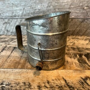 May include: Vintage metal flour sifter with a handle and a wooden knob. The sifter has a cylindrical shape with horizontal bands and a handle on the side. The metal has a distressed, aged appearance, suggesting it is an antique kitchen tool.