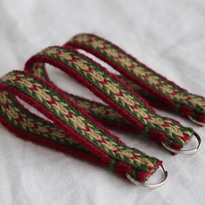 Könnte beinhalten: Drei gewebte Schlüsselanhänger mit einem geometrischen Muster in Rot, Grün und Beige. Jeder Schlüsselanhänger hat eine Schlaufe und einen silberfarbenen Metallring. Die Schlüsselanhänger liegen auf einer weißen Oberfläche.