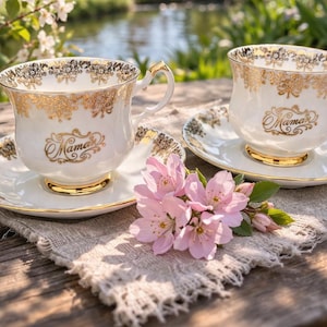 Peut inclure: Deux tasses et soucoupes blanches avec des bordures dorées et le mot "Mama" en lettres dorées. Les tasses et les soucoupes sont posées sur une table en bois avec une fleur rose et une serviette en lin. L'arrière-plan comprend un étang et des arbres en fleurs.