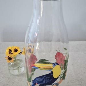 Peut inclure: Bouteille à lait en verre transparent avec un couvercle vert, ornée d'un toucan coloré. Le toucan est perché sur une branche avec des fleurs rouges. Un petit vase avec des fleurs jaunes est à gauche.