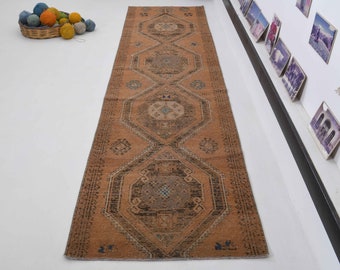Può includere: Un tappeto da runner lungo e stretto con un motivo geometrico in tonalità di marrone, beige e blu. Il tappeto ha un aspetto usurato e vintage.