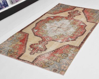 Puede incluir: Una alfombra turca vintage con fondo beige y un medallón central en tonos rojos, rosas y azules. La alfombra tiene un aspecto descolorido y desgastado y presenta intrincados patrones geométricos.