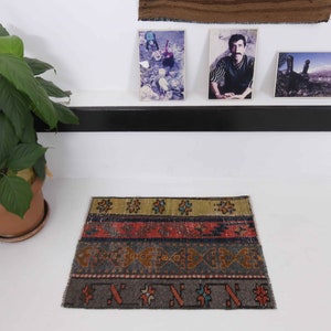 May include: A small, rectangular rug with a distressed, vintage look. The rug features horizontal bands of color including olive green, red, and gray, with geometric and floral patterns. The rug is placed on a white surface.