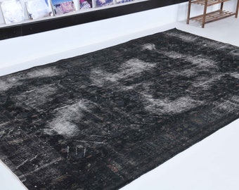 May include: A large, rectangular, distressed black rug with faded white and gray accents. The rug has a vintage, worn appearance, with visible patterns and textures. The rug is placed on a white surface.