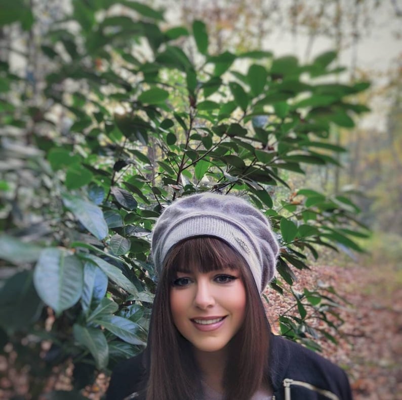 May include: A woman with long brown hair wearing a gray beret with a silver embellishment. She is smiling and looking at the camera.