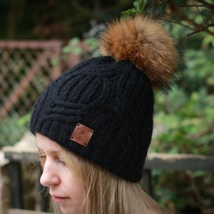 May include: A black knit beanie featuring a textured cable pattern and a large, fluffy brown faux fur pom-pom on top. A small, square, tan-colored leather-like patch is stitched onto the front cuff, displaying a circular embossed logo design.