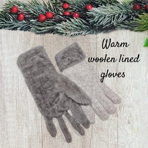 Op de afbeelding: Een paar grijze, pluche handschoenen met een grijze wollen voering. De handschoenen liggen op een houten oppervlak met een takje groen met rode bessen op de achtergrond. De tekst "Warm woolen lined gloves" is in wit op een zwarte achtergrond geschreven.