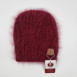 Op de afbeelding: Een bordeauxrode, pluche gebreide beanie met een bruin lederen label waarop staat "New Trend Studio" en "Kasjmier 100% natuurlijk en handgemaakt Exclusief ontwerp Gemaakt in Italië".