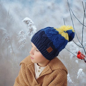 May include: A child wearing a blue and yellow knitted hat with a pom-pom on top. The hat has a cable knit design and a brown leather label. The child is wearing a brown sweater and is standing in a snowy setting.
