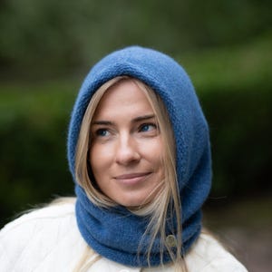 Könnte beinhalten: Eine blaue Strickkapuze mit einer weichen, strukturierten Oberfläche, die das Gesicht umrahmt. Die Kapuze verfügt über ein kleines, kreisförmiges Metallschild an der unteren Seite, das ein dezentes Detail zum einfarbigen Strickstoff bildet.